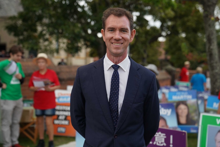 Sam Crosby stands smiling, looking at the camera, in front of campaign posters and party volunteers.