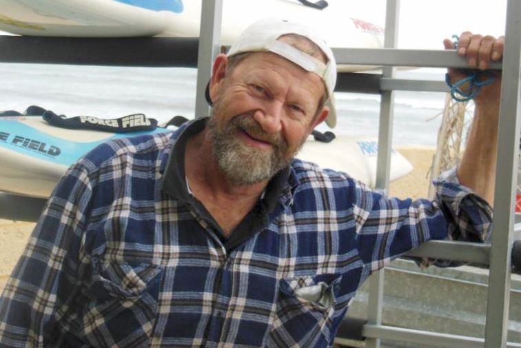 Jeffrey Doyle smiles for a photo wearing a blue flannel shirt and white cap backwards standing with a vehicle on the beach.