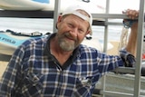 Jeffrey Doyle smiles for a photo wearing a blue flannel shirt and white cap backwards standing with a vehicle on the beach.