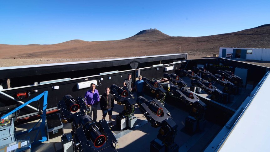 A team of astronomers stand next to telescopes at a facility in the desert.