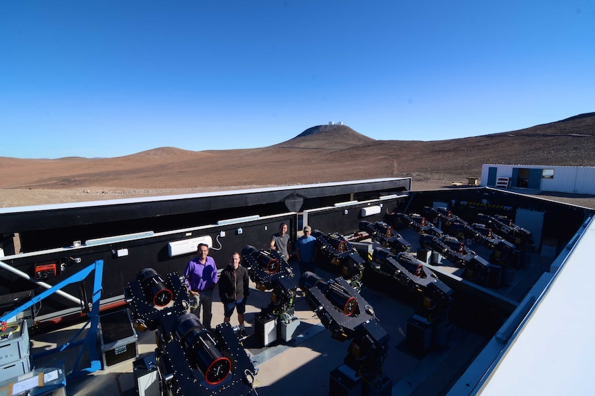 A team of astronomers stand next to telescopes at a facility in the desert.