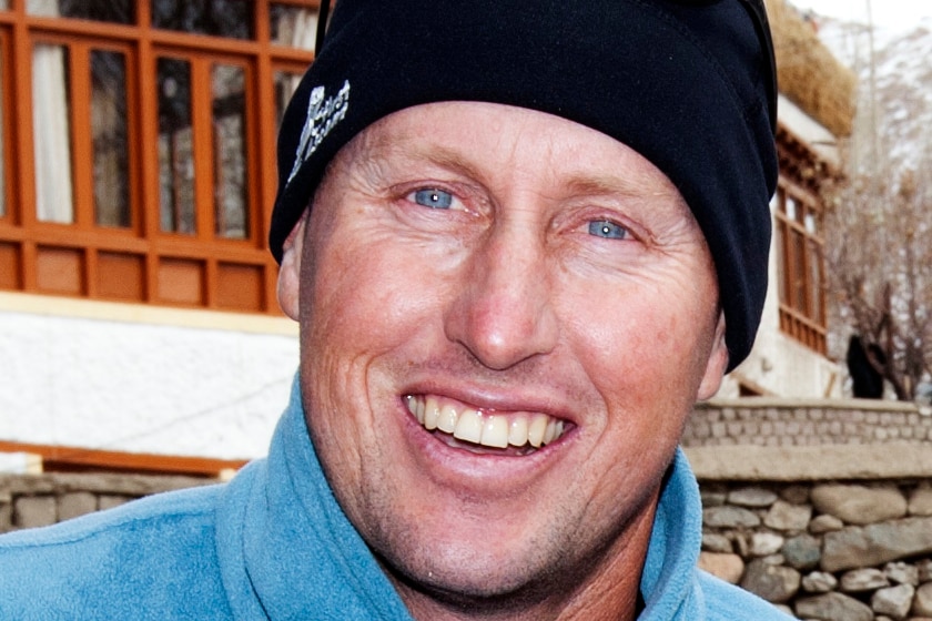 A man smiles, wearing a black beanie, sunglasses on his head and a blue jumper.