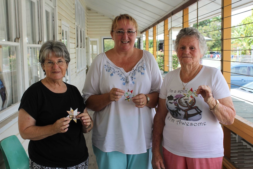 Three ladies stand on a verandah handing woven paper stars