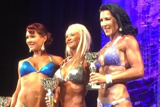 Three women in bikinis pose with trophies