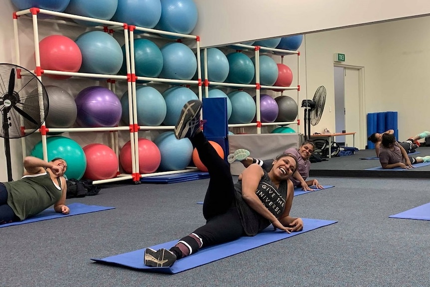 Three women are lying on mats on the ground, laughing with their legs in the air. There is a shelf of exercise balls behind them