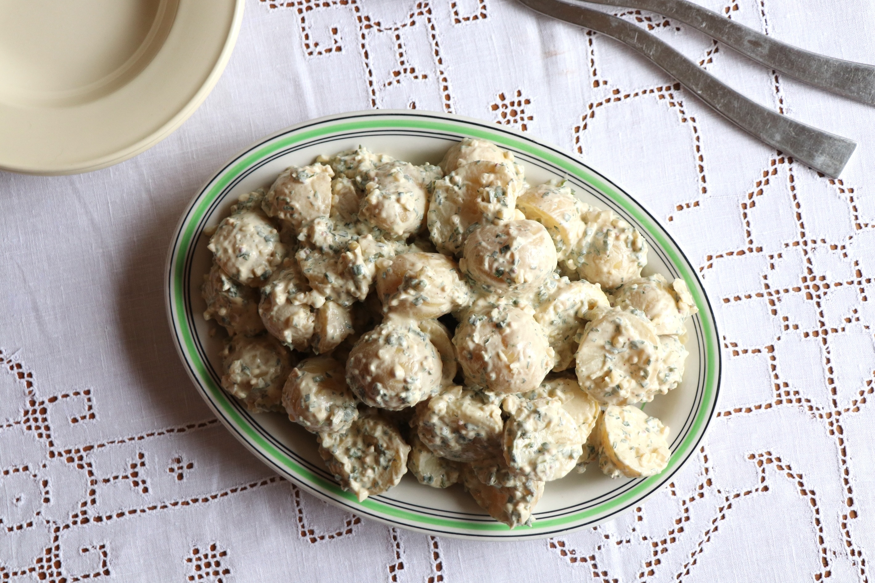 Un plato ovalado de ensalada de patatas sobre una mesa con un mantel blanco. Un plato y cubiertos para servir cerca.
