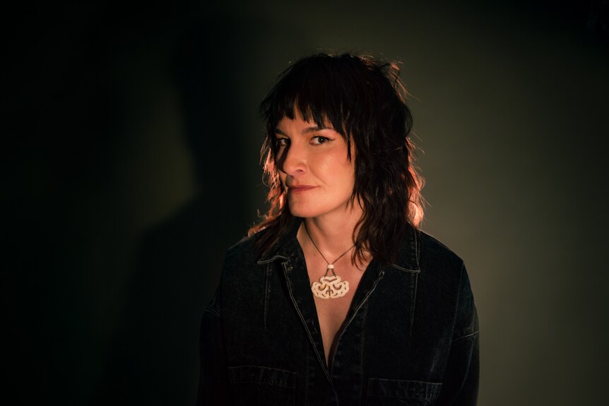 Jen Cloher wearing a dark denim shirt and large necklace looks to the camera from the corner of her eyes