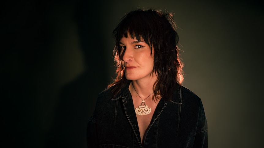 Jen Cloher wearing a dark denim shirt and large necklace looks to the camera from the corner of her eyes