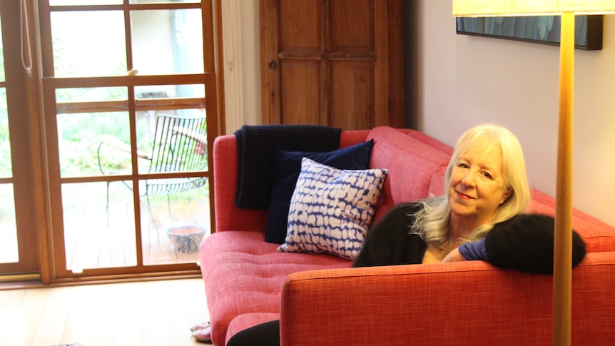 Donna Ward with shoulder-length blonde hair sits on a large red couch under a lamp smiling slightly.