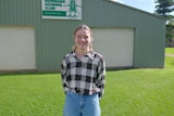 A smiling teen with brown hair tied back, wears a checked top, blue jeans. Stands on grass in front of shed.