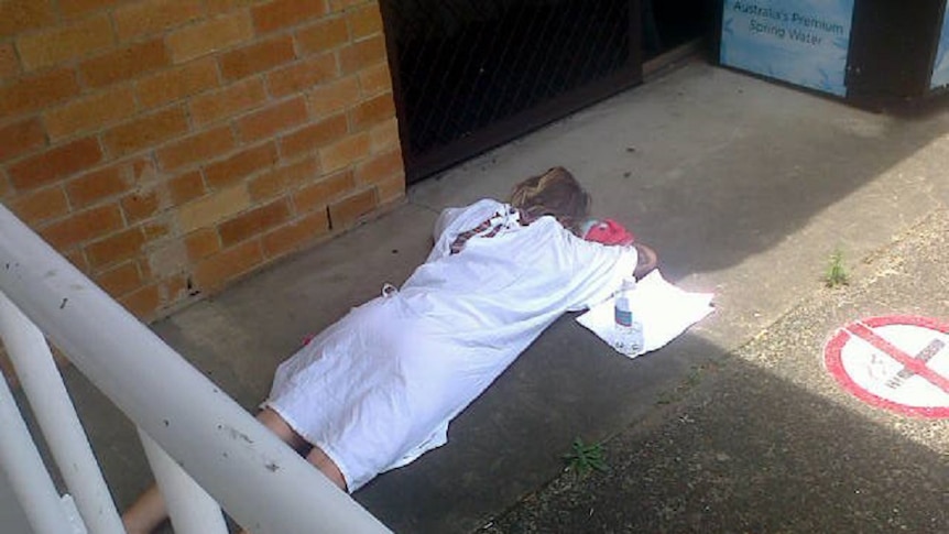 A woman in a hospital gown lies on concrete outside the Ballina Emergency Department.