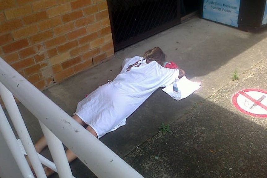 A woman in a hospital gown lies on concrete outside the Ballina Emergency Department.