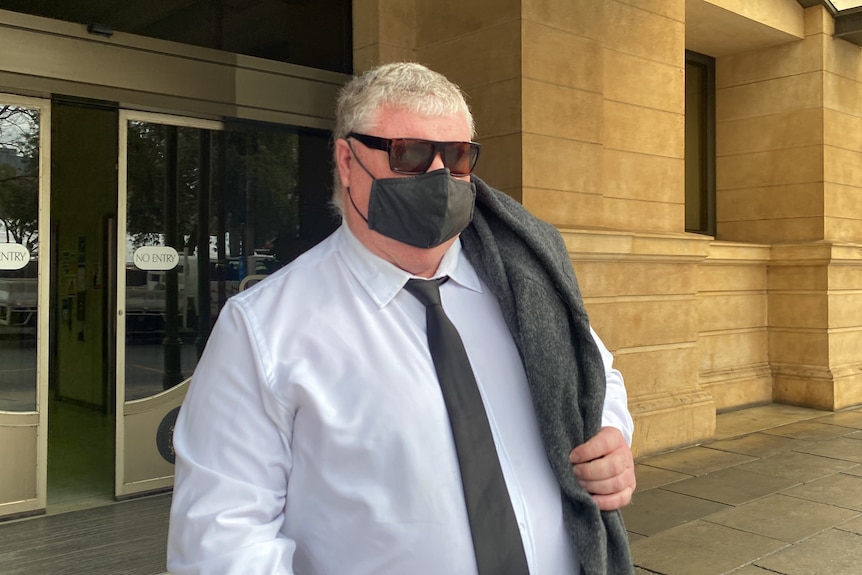 A man wearing a shirt, tie and face mask with a jumper over his shoulder walks outside court building