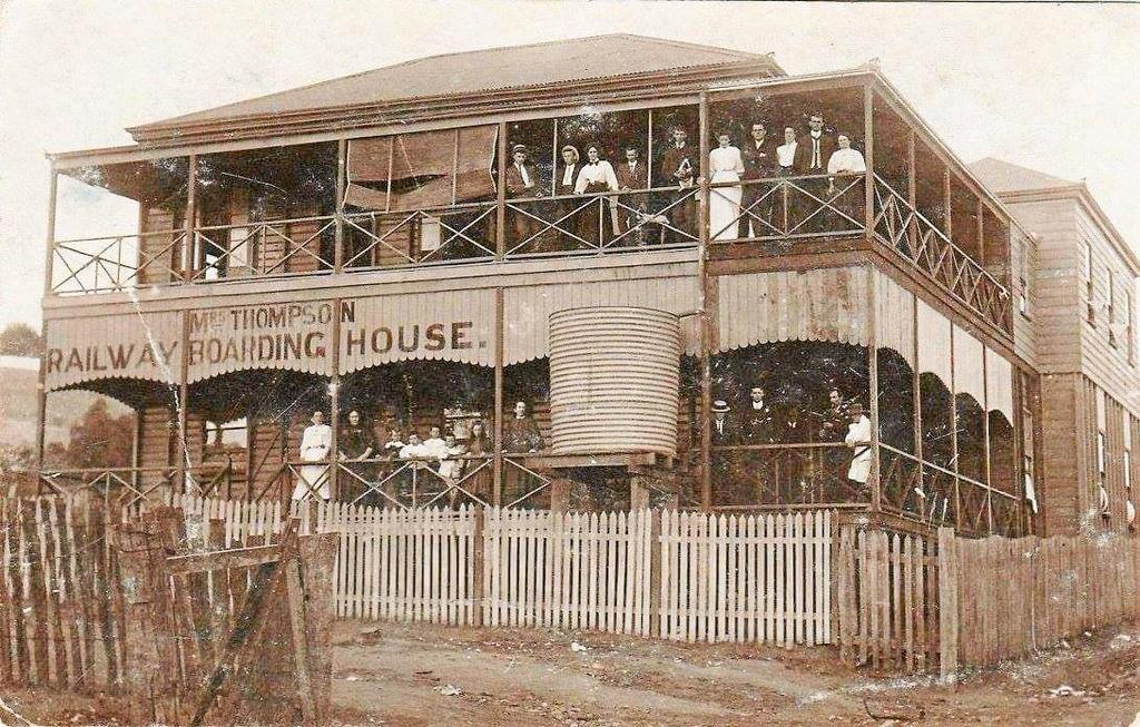 A old sepia photo of a railway boarding house with people standing on two storey verandahs. 