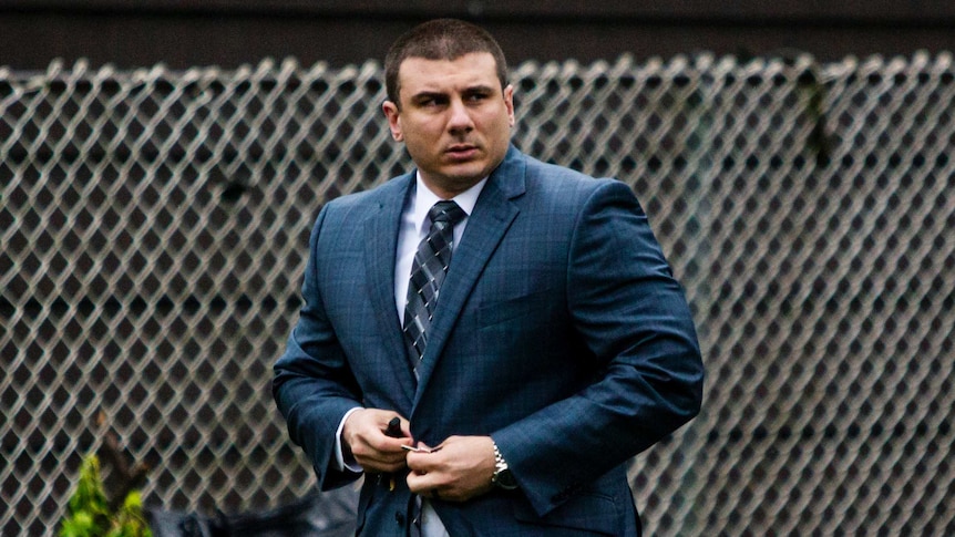 A white man buttons up the top of his teal check suit in front of a silver wire fence.