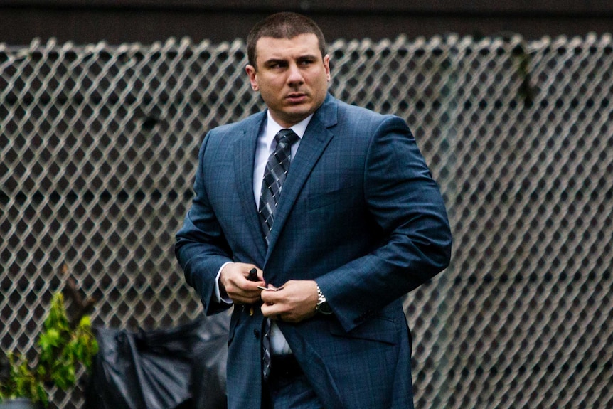 A white man buttons up the top of his teal check suit in front of a silver wire fence.