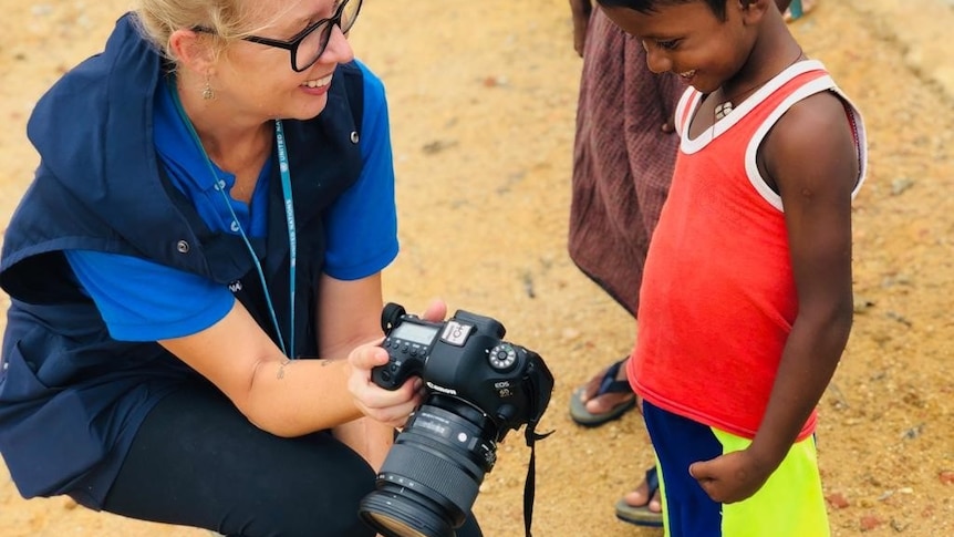 A western woman kneels down next to a young Burmese boy and shows him photographs in her camera.