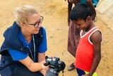 A western woman kneels down next to a young Burmese boy and shows him photographs in her camera.