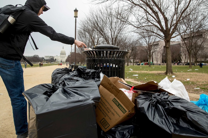 A man throws rubbish into a bin that is surrounded by garbage bags.