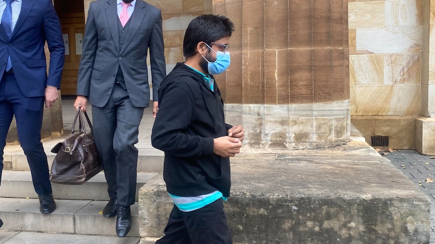 A man in a dark hoodie, glasses and face mask walks next to two men in business suits outside the court building