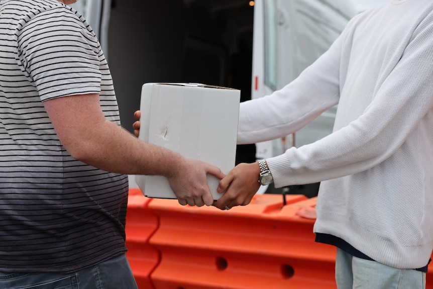 Two males passing a white box to each other, with orange barricade behind