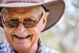A close-up of a man smiling as he stands in the wilderness. He's wearing a wide-brimmed hat and sunglasses