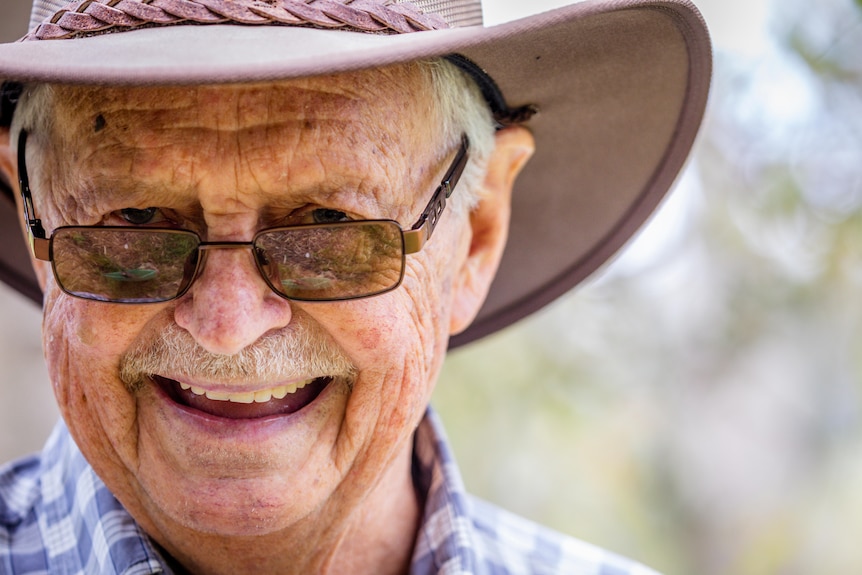 A close-up of a man smiling as he stands in the wilderness. He's wearing a wide-brimmed hat and sunglasses