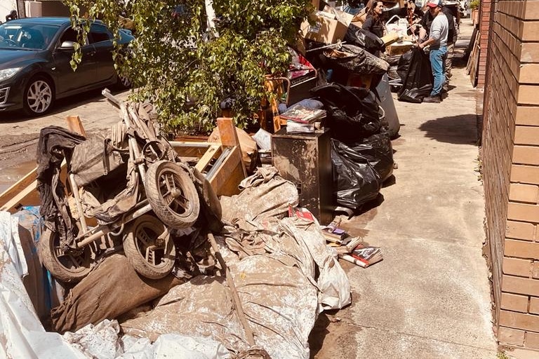 A pile of flood-damaged items including furniture and a walker piled along the side of a concrete pathway