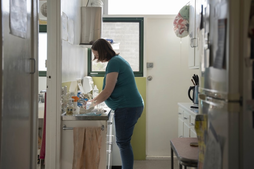 A young pregnant woman with medium length brown hair. She is in the kitchen, washing dishes