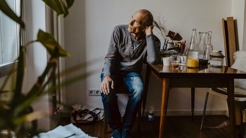 Man staring out a window with a sad look on his face sitting at a table with glasses and bottles on it