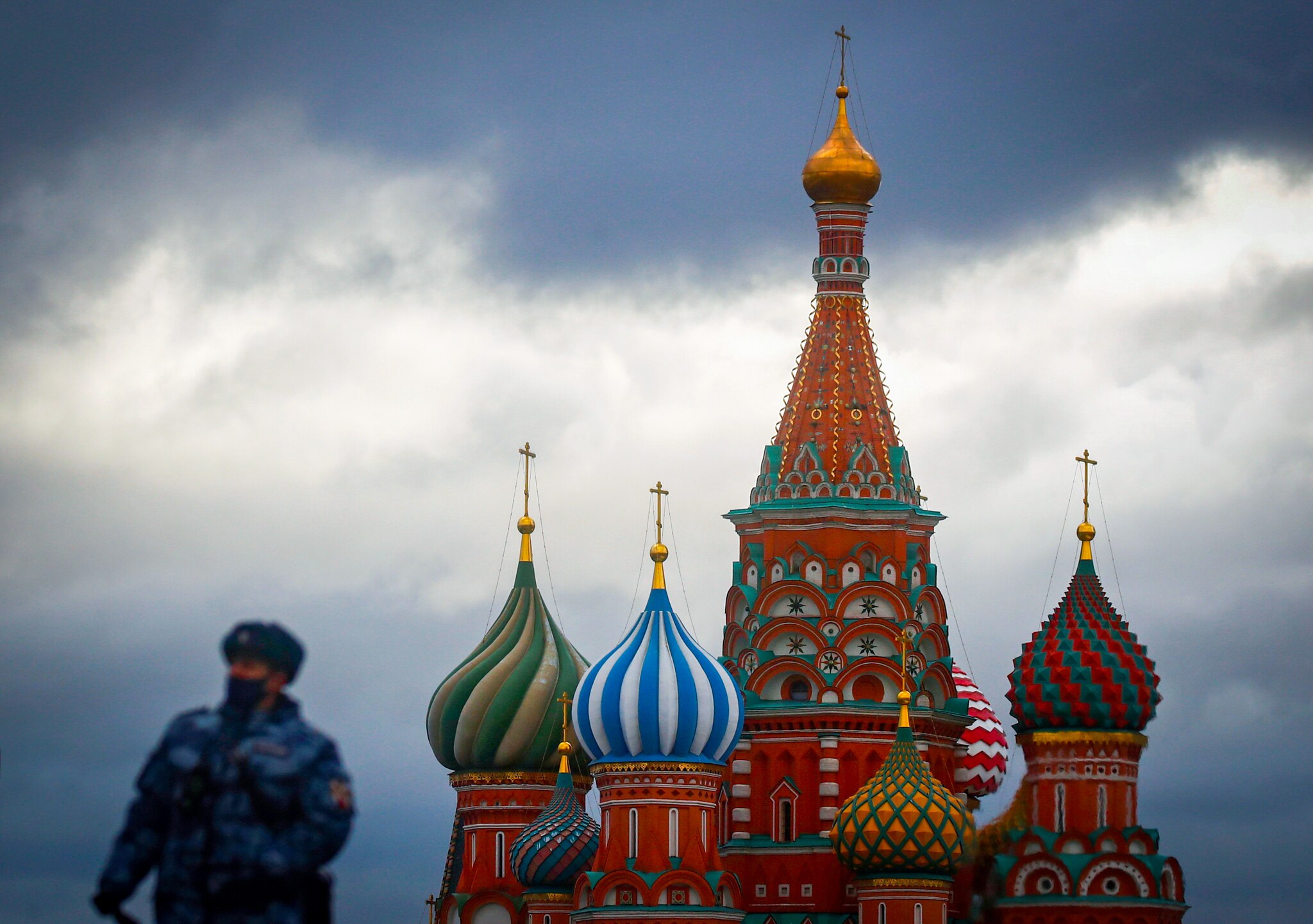A masked soldier, out of focus, next to the St Basil's Cathedral, a colourful building with curves turrets, against a grey sky