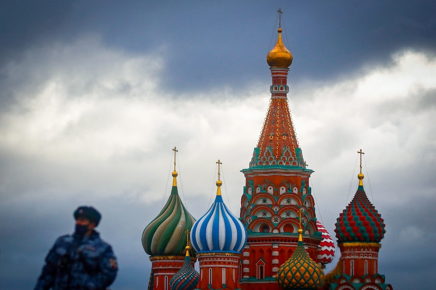 A masked soldier, out of focus, next to the St Basil's Cathedral, a colourful building with curves turrets, against a grey sky