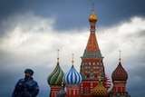 A masked soldier, out of focus, next to the St Basil's Cathedral, a colourful building with curves turrets, against a grey sky