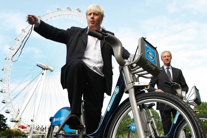 Looking up, Boris Johnson poses on a London share bike with the London eye behind him.