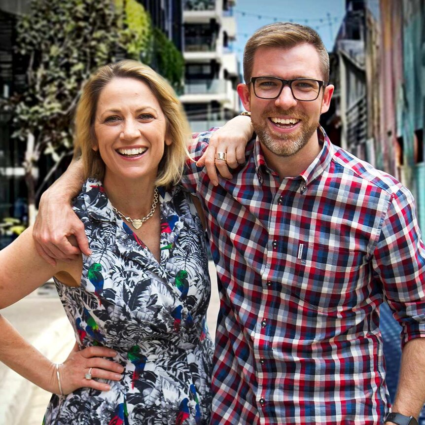 A blonde woman and a man wearing glasses standing arm in arm in a city alleyway.