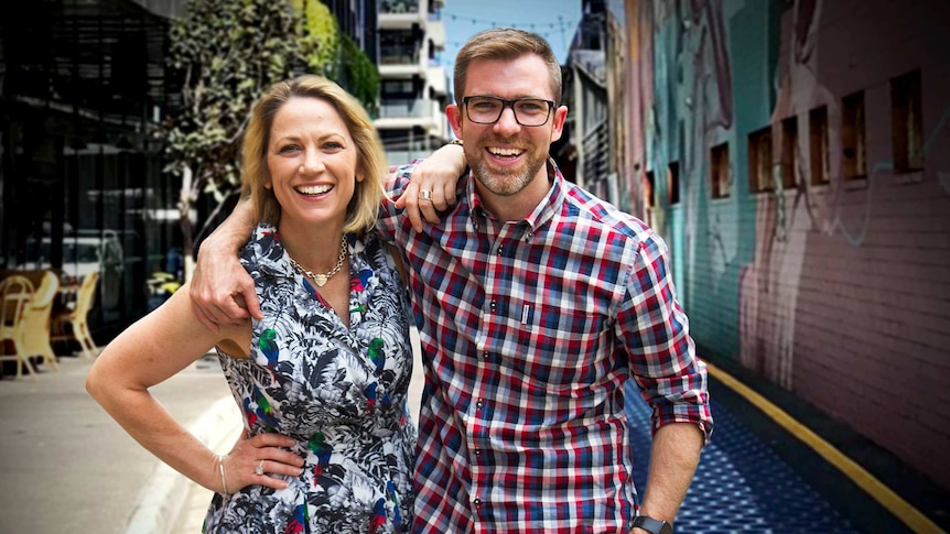 A blonde woman and a man wearing glasses standing arm in arm in a city alleyway.