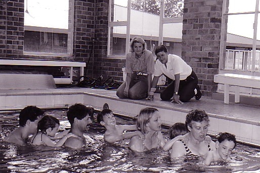 Men and women hold children in a swimming pool in a black and white photo.