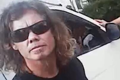 A CCTV frame of a man with shoulder length hair, wearing sunglasses and a black t-shirt, next to car.