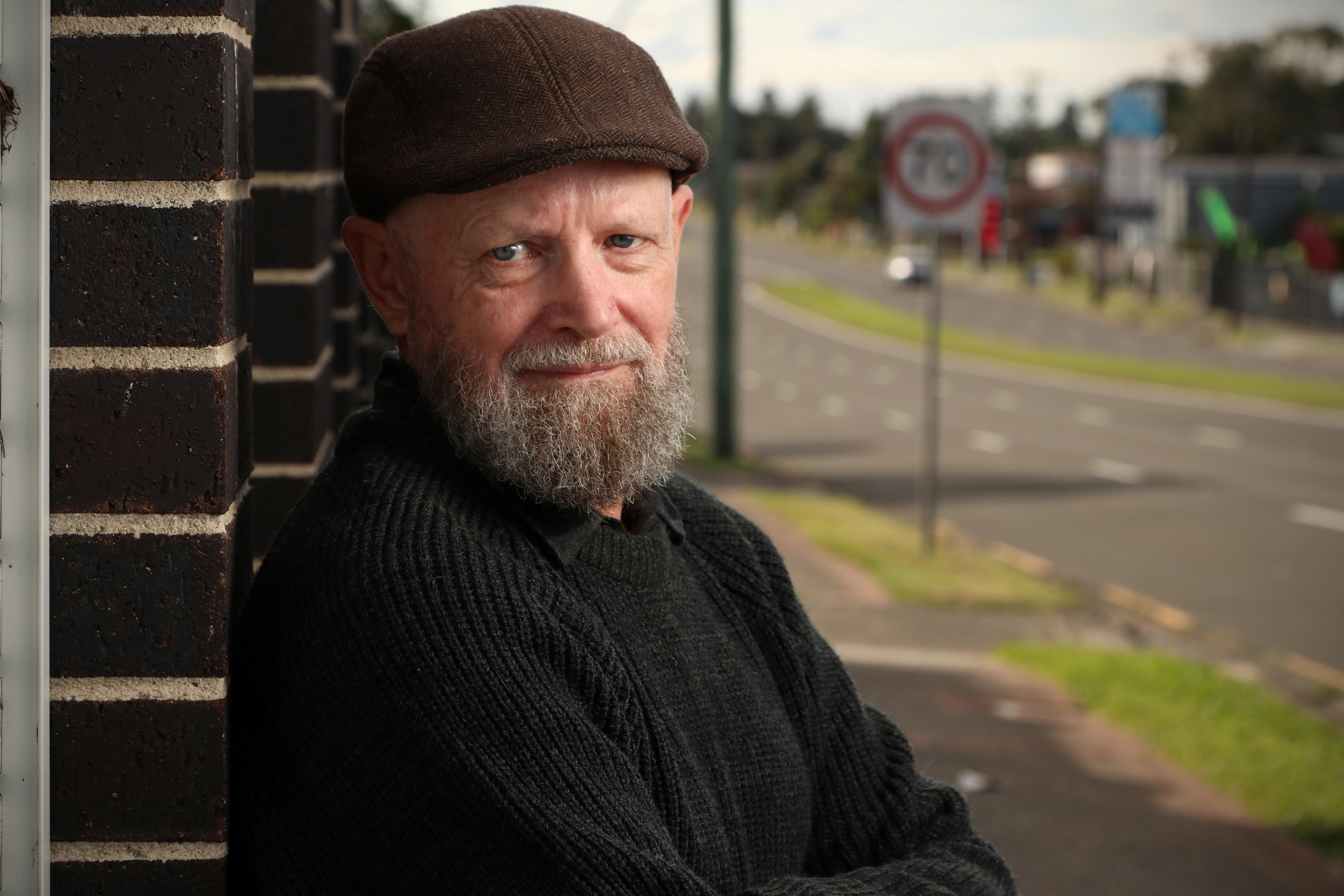A bearded, middle-aged man in a flat cap leans against a brick wall near a main road.