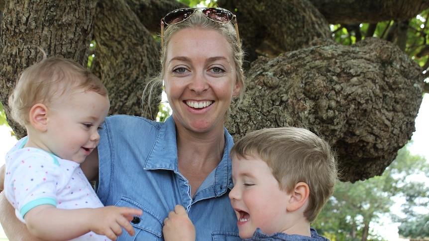 young woman stands holding two young children who are interacting with each other and mum is smiling looking at the camera