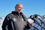 A bald man wearing black jacket with ITF logo, stands and talks into a microphone outside, blue sky.