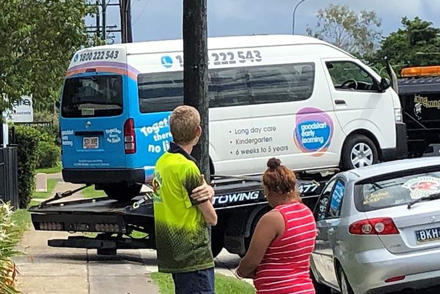 A man and a woman watch a minibus being towed away