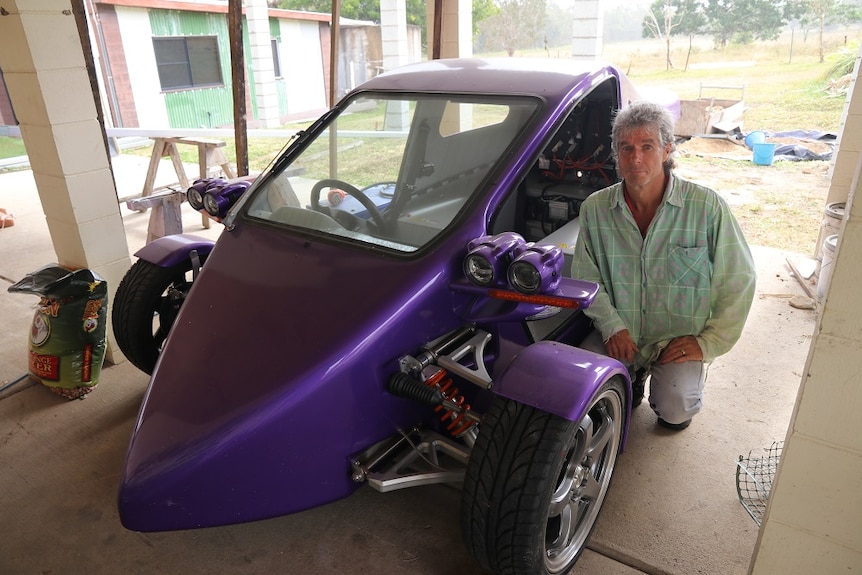A man crouches next to a purple three-wheeled car/motorcycle