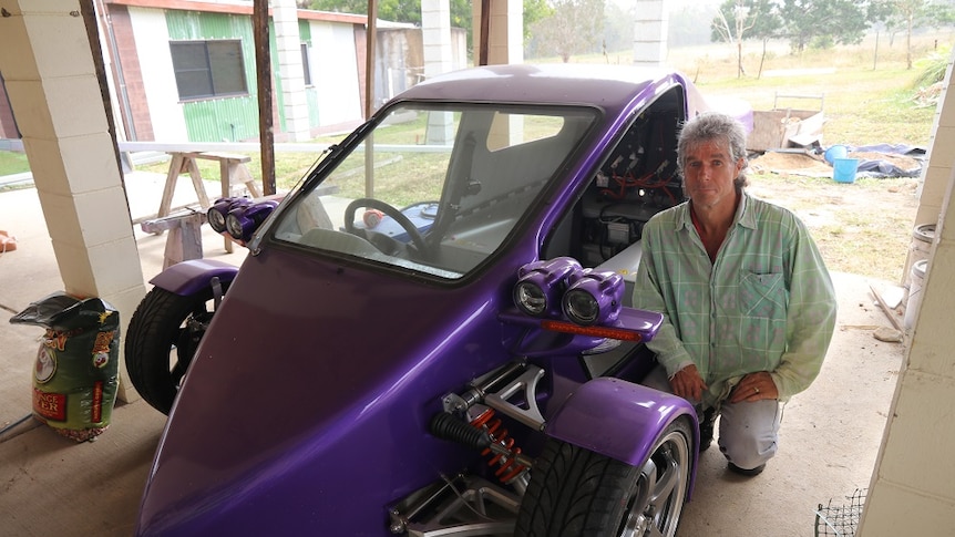 A man crouches next to a purple three-wheeled car/motorcycle
