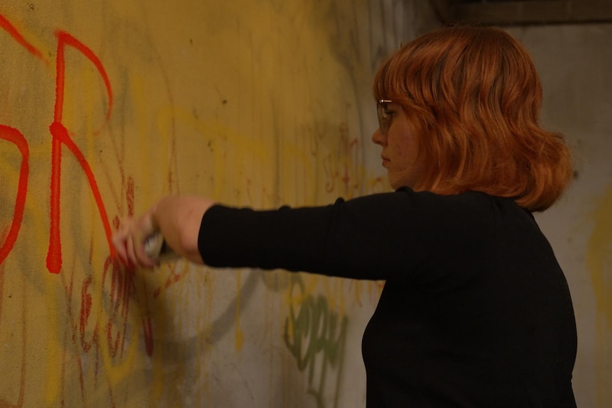A young woman with red hair sprays the letters 'SR' in red on a wall.