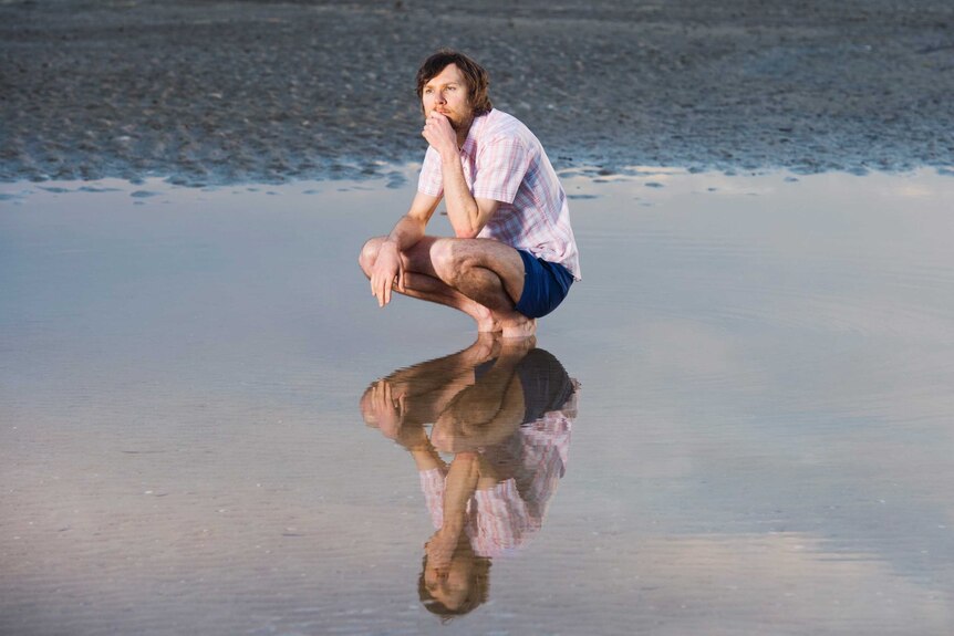A man with a beard squats down in a pool of water, gazing wistfully into the distance. He is reflected in the water.