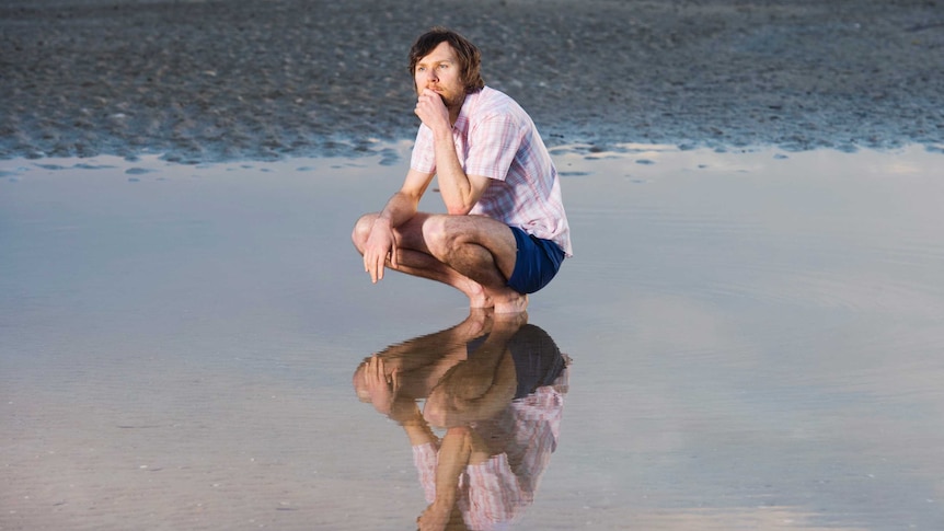 A man with a beard squats down in a pool of water, gazing wistfully into the distance. He is reflected in the water.