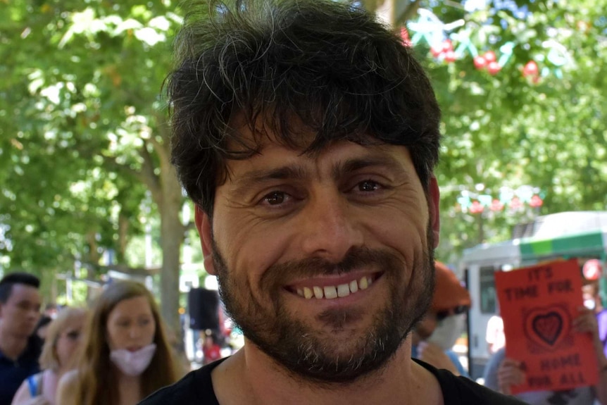 A man smiling while standing outdoors under trees.