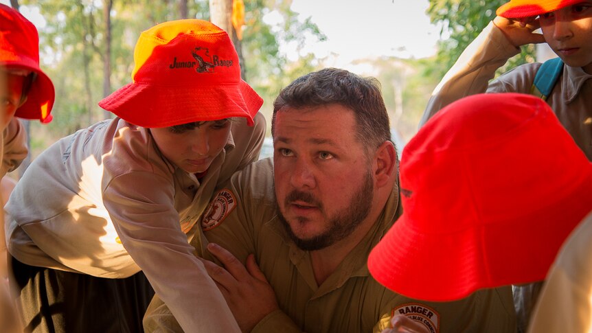 A Northern Territory parks and wildlife ranger dressed in a khaki uniform is surrounded by children wearing bright orange hats.
