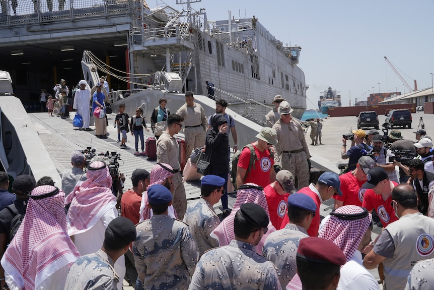 Many people and uniformed officers for the US Navy walk down a large ramp from a ship to the dock.